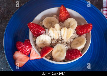 Haferflocken in einer Schüssel auf einem blauen Teller, serviert mit Erdbeeren und Bananen in Herzform geschnitten, gesundes Frühstück, gesunde Ernährung, Stockfoto