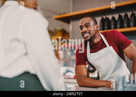 Ein fröhlicher männlicher Barista auf einer Schürze bietet ausgezeichneten Kundenservice in einem modernen Café. Die helle Inneneinrichtung und die angenehme Interaktion zeigen ein warmes, Stockfoto