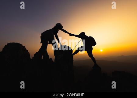 Silhouette von Menschen, die sich gegenseitig helfen, bei Sonnenaufgang einen Berg hinauf zu wandern. Eine helfende Hand und ein Lifestyle-Konzept mit aktiver Passform. Hilfe und Erfolg Stockfoto