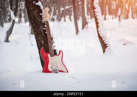 Rote E-Gitarre im verschneiten Wald. Fender Squire Stratocaster lehnte sich an einen Baumstamm im Winterwald, unterer Teil der Gitarre im Schnee. Stockfoto