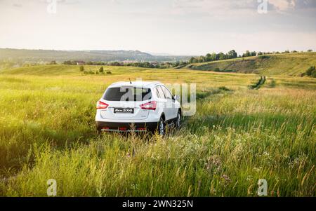 Weißer Suzuki SX4 in einer hügeligen Landschaft. Rückansicht der kompakten Überkreuzung auf einer grünen Wiese - Wochenende weg von der Stadt Stockfoto