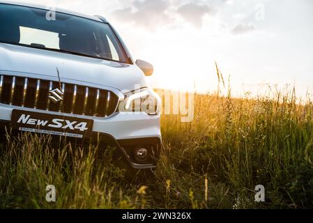 Weißer Suzuki SX4 in einem hügeligen Feld bei Sonnenuntergang. Nahaufnahme der Vorderseite der kompakten Überkreuzung auf einer Wiese - Wochenende weg von der Stadt Stockfoto