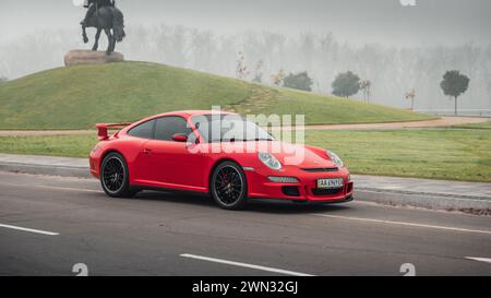 Roter Porsche 911 an einem nebeligen Morgen. Dreiviertelansicht des Sportwagens Porsche 997 vor dem Pferdedenkmal auf einem Hügel. Stockfoto