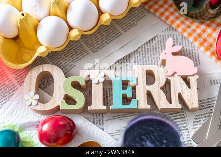 Augsburg, Bayern, Deutschland - 28. Februar 2024: Ostern, Holzschriftzug auf einem Tisch mit Zeitung und Zubehör zum Färben von Ostereiern *** Ostern, Schriftzug aus Holz auf einem Tisch mit Zeitung ausgelegt und Zubehör zum Ostereier färben Stockfoto