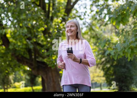 Eine ältere Frau mit grauen Haaren lächelt, während sie Musik hört und in einem lebhaften grünen Park joggt. Stockfoto