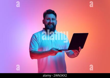 Hübscher junger Mann lächelt in die Kamera und hält den Laptop im Neonlicht vor lebendigem Hintergrund im Studio mit Farbverlauf. Stockfoto