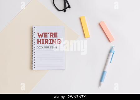 Ein weißer Schreibtisch mit einem Notizbuch mit „We Are Hiring“-Schriftzug, blauem Stift, Textmarkern, die Arbeitsmöglichkeiten und die Arbeitsumgebung symbolisieren Stockfoto