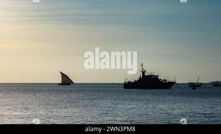 Ein traditionelles Dhow-Segelboot nähert sich in der Abenddämmerung einem tansanianischen Marinekatrouillenboot in Stone Town, Sansibar, Tansania Stockfoto