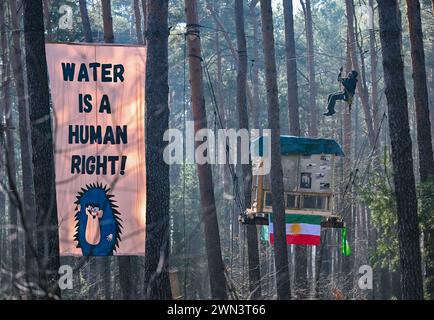 29. Februar 2024, Brandenburg, Grünheide: Zwischen Bäumen hängt ein Aktivist der Initiative „Stop Tesla“ neben einem Banner mit der Aufschrift „Wasser ist ein Menschenrecht!“ In einem Kiefernwald in der Nähe der Tesla Gigafactory Berlin-Brandenburg. Rund 80 Aktivisten der Initiative „Stop Tesla“ haben ein Waldstück in der Nähe der Automobilfabrik in Grünheide in Brandenburg besetzt. Sie wollen gegen die geplante Fabrikerweiterung protestieren. Die Gruppe errichtete Baumhäuser auf dem rund 120 Hektar großen Waldstück, das für die Erweiterung des Geländes gerodet werden soll. Die Beschäftigung ist für einen in geplant Stockfoto