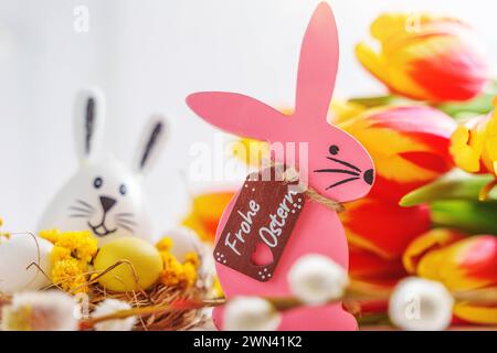 Augsburg, Bayern, Deutschland - 28. Februar 2024: Frohe Ostergrüße auf einem rosa hölzernen Osterhasen neben Blumen Dekoration *** Frohe Ostern Ostergrüße auf einem Pinken Osterhasen aus Holz neben Blumen Dekoration Stockfoto