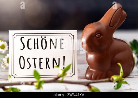 Augsburg, Bayern, Deutschland - 28. Februar 2024: Happy Easter-Schriftzug auf einer Grußkarte neben einem Schokoladen-Osterhasen. FOTOMONTAGE *** schöne Ostern Schriftzug auf einer Grußkarte neben einer Osterhase aus Schokolade. FOTOMONTAGE Stockfoto