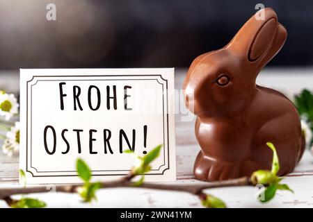 Augsburg, Bayern, Deutschland - 28. Februar 2024: Happy Easter-Schriftzug auf einer Grußkarte neben einem Schokoladen-Osterhasen. FOTOMONTAGE *** Frohe Ostern Schriftzug auf einer Grußkarte neben einer Osterhase aus Schokolade. FOTOMONTAGE Stockfoto