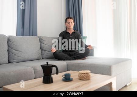 Frau, die auf einer Couch sitzt und mit geschlossenen Augen meditiert. Relaxation, Stressentlastungskonzept. Stockfoto