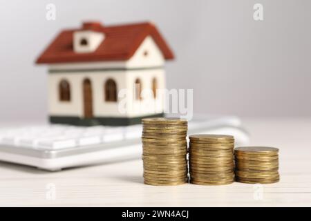 Hypothekenkonzept. Münzstapel, Modellhaus und Taschenrechner auf weißem Holztisch, selektiver Fokus Stockfoto