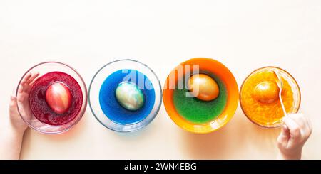 Foto von oben von einer Hand eines Mädchens Kind, das festliche Ostereier mit bunten Farben auf dem Tisch zu Hause malte und färbte. Kinder machen sich auf EA vorbereitet Stockfoto