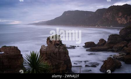 Raue Meeresküste mit Wellen und einigen Häusern und scharfen Klippen während Sonnenuntergang, Neuseeland Stockfoto