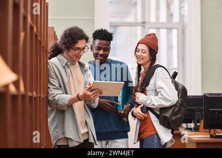 Multiethnische Gruppe von drei fröhlichen jungen Menschen, die an Regalen in der Universitätsbibliothek stehen und digitale Tablets nutzen Stockfoto