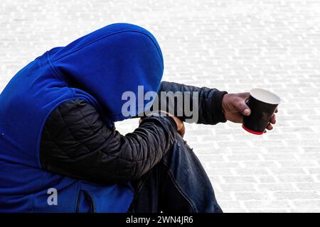 Afroamerikaner mit Jacke und königsblauem Kapuzenpulli, ausgestrecktem Arm, der eine Tasse hält und Spenden sucht. Stockfoto