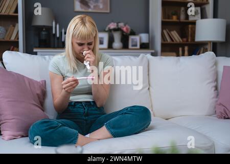 Einzelne traurige Frau, die sich über einen Schwangerschaftstest beschwert, sitzend auf einer Couch im Wohnzimmer zu Hause, gestresste Frau, positive oder negative medizinische Taten Stockfoto