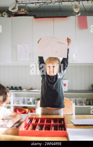 Porträt eines Mädchens, das Papier mit Zeichnung zeigt, während er in der Nähe der Bank im Klassenzimmer steht Stockfoto