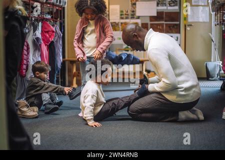 Seitenansicht eines männlichen Lehrers, der im Kindergarten mit Schuhen hilft Stockfoto