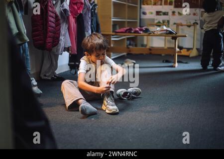 Junge, der Schnürsenkel bindet, während er auf dem Boden in der Garderobe in der Vorschule sitzt Stockfoto