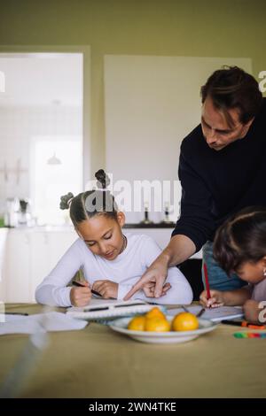 Vater hilft Töchtern, während er Hausaufgaben macht, am Esstisch sitzt Stockfoto