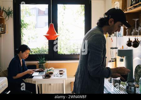 Männlicher Architekt bereitet Kaffee vor, während weibliche Kollegin ihr Smartphone im Heimbüro benutzt Stockfoto