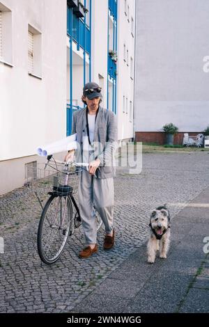Der männliche Architekt hält Bauplanerollen, die mit dem Fahrrad und mit dem Hund auf dem Fußweg laufen Stockfoto