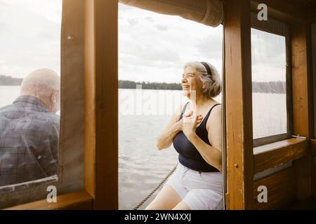 Glückliche Seniorin mit Händen auf der Brust, die neben dem Mann auf dem Hausboot sitzt Stockfoto