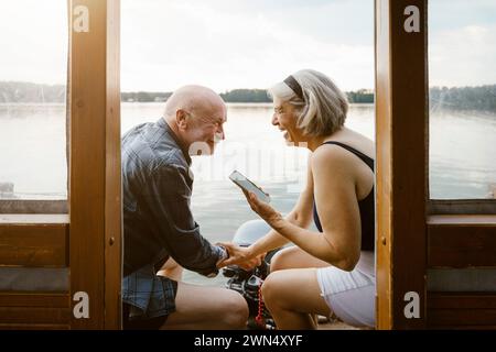 Glückliches Seniorenpaar mit Smartphone, das auf dem Hausboot sitzt Stockfoto