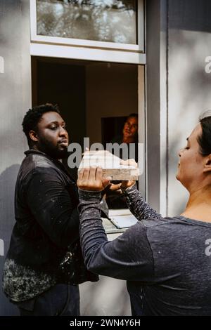Männliche und weibliche Bauarbeiter, die eine Planke durch das Fenster des Hauses führen Stockfoto