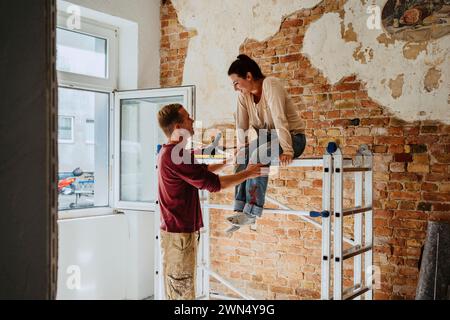 Mann, der mit einer Frau spricht, die auf einer Leiter vor einer Ziegelmauer sitzt, während der Hausrenovierung Stockfoto