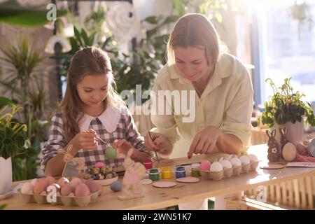 Fröhliche kaukasische Mutter und Tochter sitzen am Holztisch in der Werkstatt und dekorieren Eier für Ostern Stockfoto