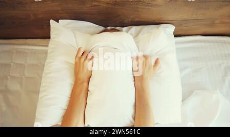 Ungesunder Schlaf, Frau, die an Schlaflosigkeit leidet, liegt auf dem Bett und hält in den Händen, die ihren Kopf mit weißem Kissen im Schlafzimmer zu Hause bedecken Stockfoto