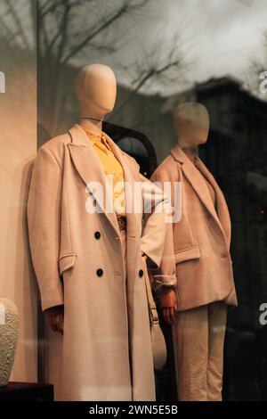 Zwei stilvolle Schaufensterpuppen in saisonalem Stil in einem Schaufenster. Beiger langer Mantel und ein hellgelbes Hemd. Stockfoto