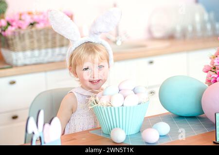 Kleines Mädchen mit weißen osterhasenohren lächelte glücklich, während er am Tisch neben einem Stapel von gefundenen bemalten ostereiern in der Küche am pessach-Tag saß Stockfoto