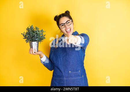 Junge kaukasische Gärtnerin hält eine Pflanze isoliert auf gelbem Hintergrund, lacht über dich, zeigt mit dem Finger auf die Kamera, mit der Hand über dem Körper, Schande Stockfoto