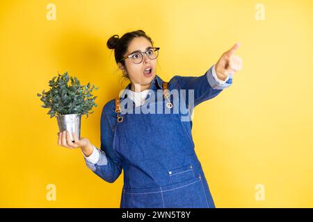 Junge kaukasische Gärtnerin, die eine Pflanze auf gelbem Hintergrund hält, mit dem Finger überrascht nach vorne zeigt, offener Mund verwundert, somet Stockfoto