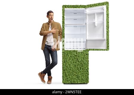 Lässiger Mann in Hemd und Jeans, lehnt sich auf einen grünen umweltfreundlichen Kühlschrank und zeigt isoliert auf weißem Hintergrund Stockfoto