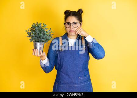 Junge kaukasische Gärtnerin, die eine Pflanze isoliert auf gelbem Hintergrund hält, depressiv und besorgt sich um Not, weinend und ängstlich. Trauriger Ausdruck Stockfoto