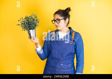 Junge kaukasische Gärtnerin, die eine Pflanze isoliert auf gelbem Hintergrund hält, entspannt mit ernstem Gesichtsausdruck. Schlicht und natürlich aussehend Stockfoto