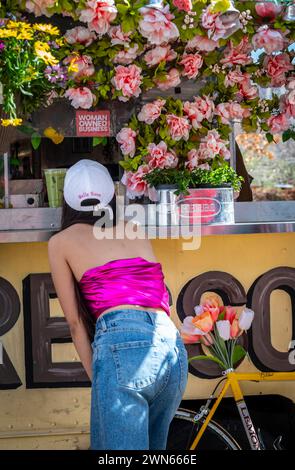 Rückansicht einer Frau im pinkfarbenen Bandeau, die Saft aus dem Food Truck bestellt hat, mit übermäßig floralem Thema und Schild, auf dem man GESCHÄFTE VON FRAUEN lesen kann Stockfoto