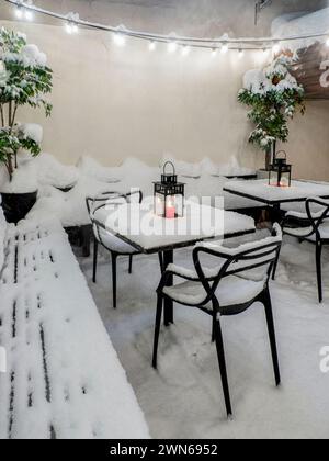 Tische und Stühle im Winter mit frischem Schnee bedeckt, brennende rote Kerze auf dem Tisch bedeckt mit frischem Schnee Stockfoto