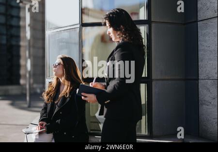 Zwei junge Frauen in Geschäftskleidung, die vor städtischem Hintergrund produktiv diskutiert werden, symbolisieren Teamarbeit und Unternehmergeist. Stockfoto