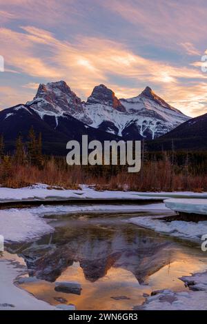 Goldener Winteruntergang bei den Three Sisters, einem Dreigestirn von Gipfeln in der Nähe von Canmore, Alberta, Kanada, mit alpenglow auf den Bergen, die sich im Fo vom Eis spiegeln Stockfoto