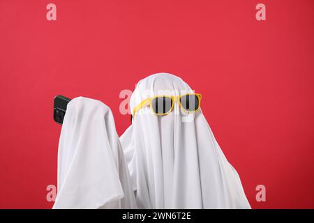 Person im Geisterkostüm und Sonnenbrille mit Smartphone auf rotem Hintergrund Stockfoto