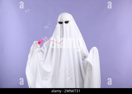 Lustiger Geist. Person in weißem Blatt und Sonnenbrille, die Seifenblasen auf violettem Hintergrund bläst Stockfoto