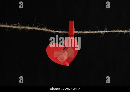 Gebrochenes Herz. Gerissenes rotes Papierherz mit medizinischen Haftverbänden auf schwarzem Hintergrund Stockfoto