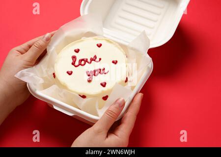 Frau, die eine Kiste mit bento-Kuchen am roten Tisch hält, Nahaufnahme. St. Valentinstag-Überraschung Stockfoto